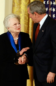 Olivia de Havilland shakes hands with President George W. Bush in a formal setting with US flag visible